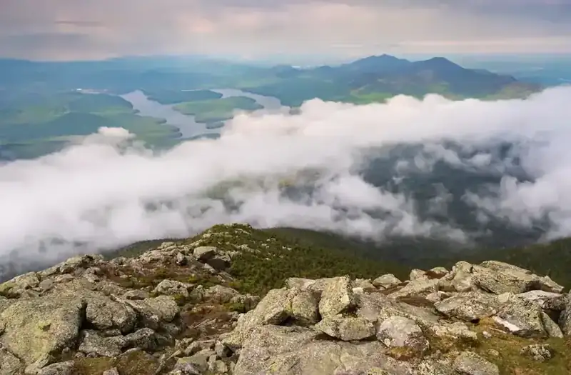 A view from a rocky Adirondack summit over Lake Placid and forested peaks, with clouds drifting below the ridge
