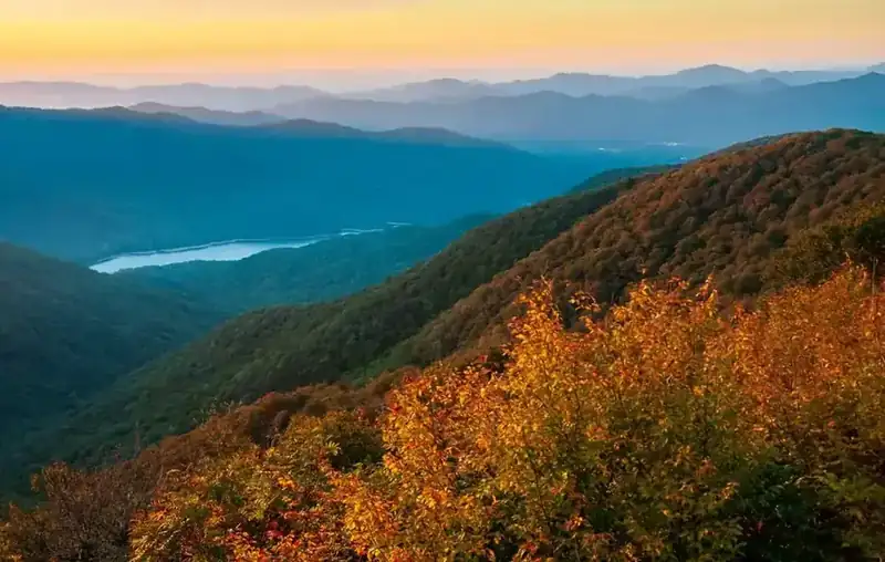 Layers of blue ridges at sunset above a reservoir, with autumn foliage in the foreground