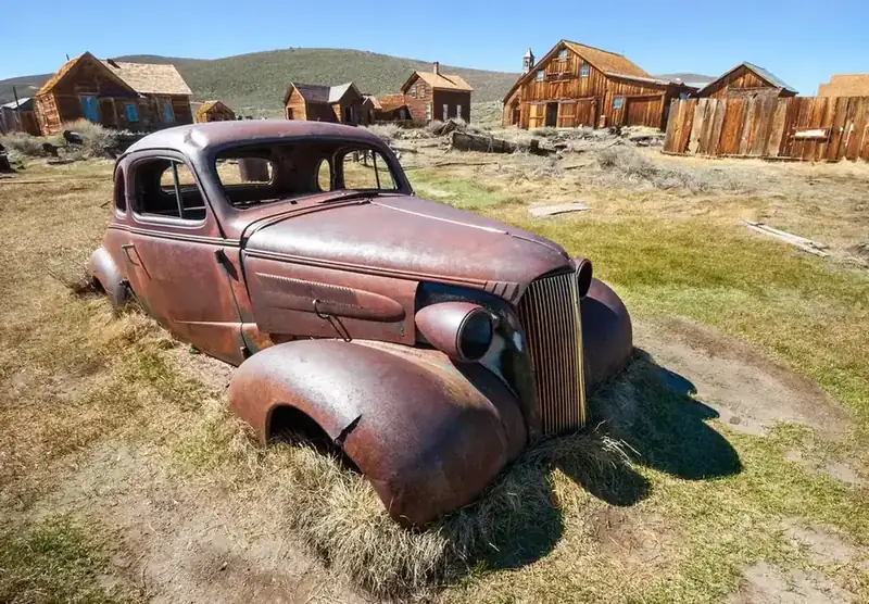 A rusting 1930s coupe sitting in the grass among the weathered buildings of Bodie